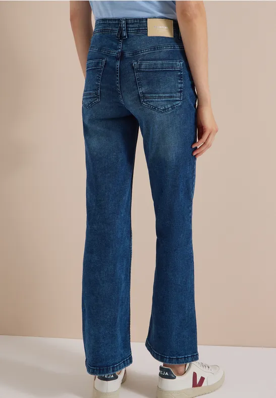 Frau steht von hinten, trägt dunkelblaue High-Waist-Jeans und weiße Sneaker vor beigeem Hintergrund.