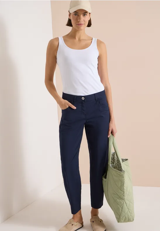 Femme en débardeur blanc et pantalon bleu marine tenant un sac matelassé vert devant un fond épuré.