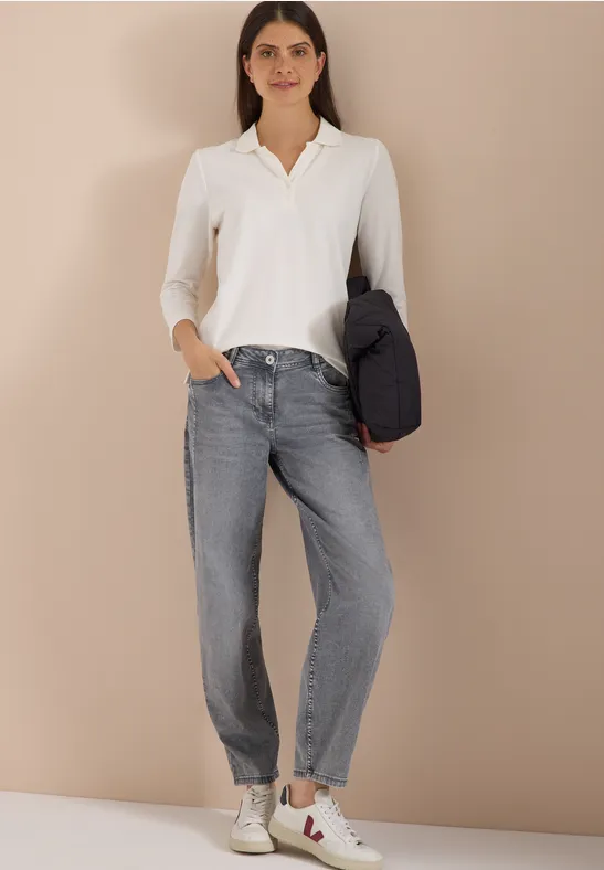 Junge Frau in weißem Hemd, grauer Jeans und Sneakers hält eine Jacke lässig unter dem Arm.