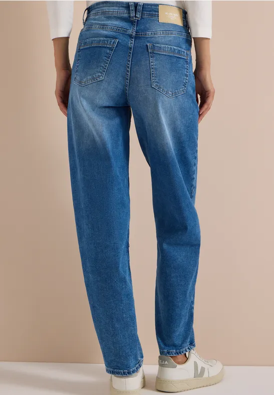 Vue de dos d’une personne portant un jean bleu ample et des baskets blanches sur fond beige.
