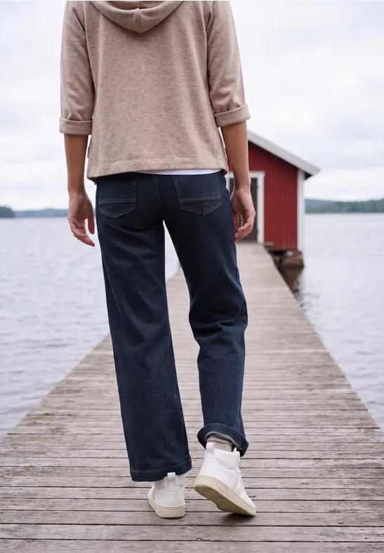 Person im Hoodie und Jeans läuft auf einem Holzsteg zum roten Bootshaus an einem ruhigen See.