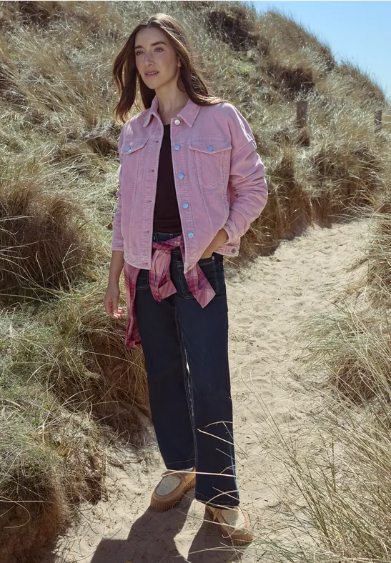 Frau in rosa Jeansjacke, weitem Dunkelblau-Jeans und beigen Sandalen läuft über einen grasigen Dünenpfad.