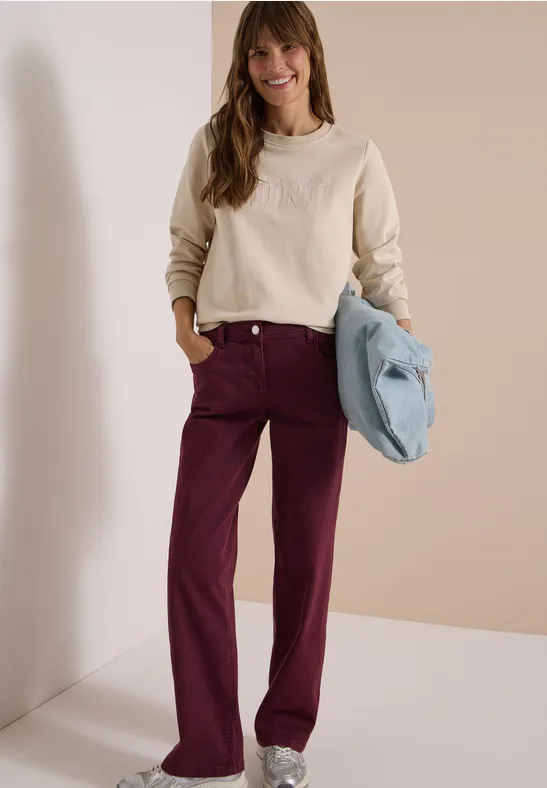 Femme debout sur fond neutre, porte un sweat beige, un pantalon bordeaux et tient un sac bleu clair.