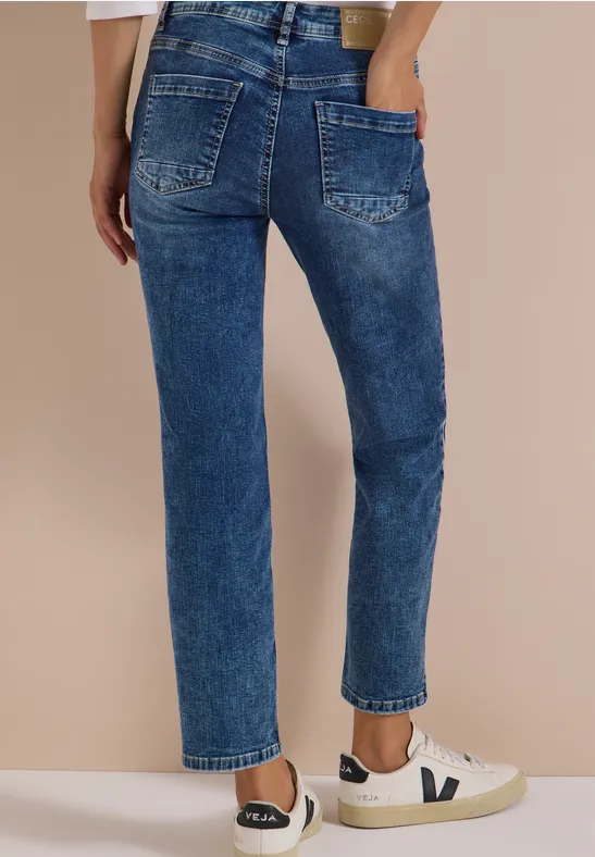 Femme en jean bleu taille haute, coupe droite, baskets blanches, sur fond beige.