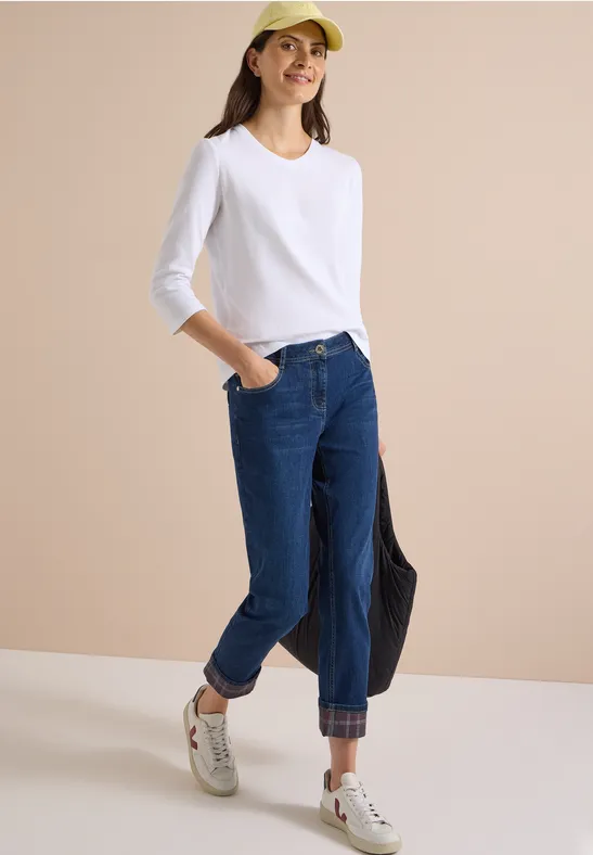 Femme en T-shirt blanc à manches longues, jean bleu, baskets blanches et casquette jaune, tenant un sac noir devant un mur beige