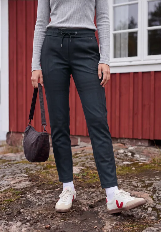 Person steht vor rotem Haus, schwarze Hose, weiße Sneaker mit rotem V, hält Tasche.