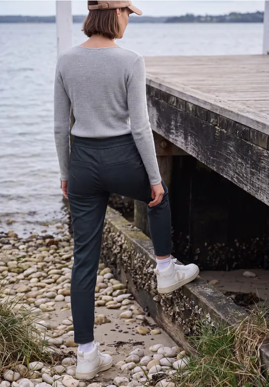 Person in grauem Oberteil, dunkler Hose und weißen Boots steht am Wasser neben einem Holzsteg.