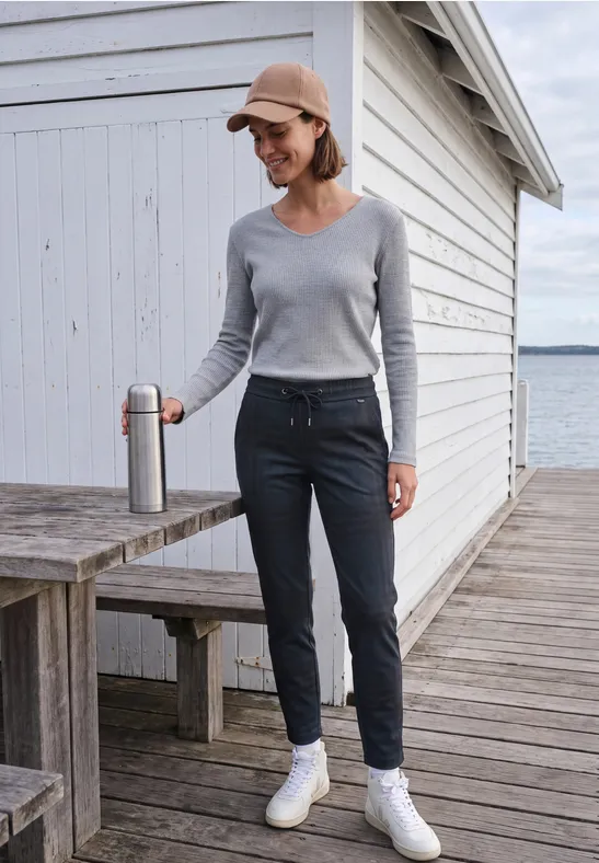Frau in grauem Pullover, dunkler Hose und Cap steht auf einem Steg vor einem Schuppen am Wasser und hält einen Becher.