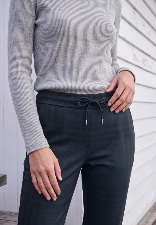 Frau in grauem Pulli und dunklen, taillierten Hosen mit Kordel vor weiß vertäfelter Wand.