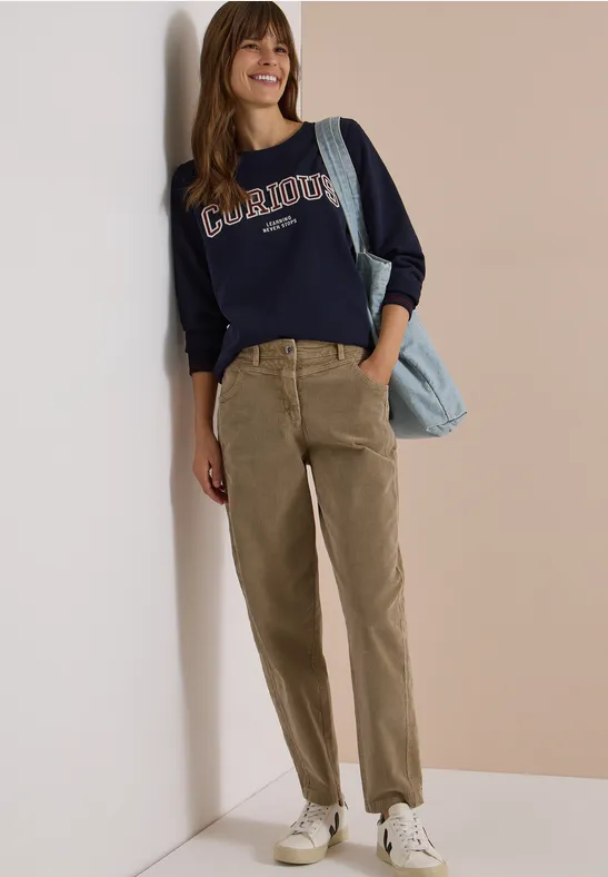 Frau lehnt an Wand, lächelt, trägt dunkelblaues Sweatshirt, olivgrüne Cordhose, weiße Sneaker und blaue Tasche.