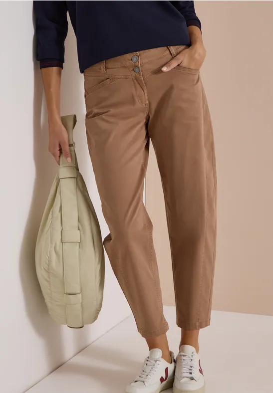Eine Frau trägt bequeme, hellbraune Hose, weiße Sneaker und dunkles Shirt, dazu eine große hellgrüne Tasche.
