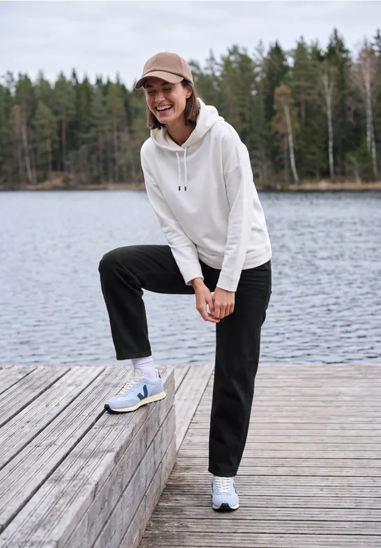Lächelnde Frau in weißem Hoodie und schwarzer Hose auf einem Steg am See, im Hintergrund Wald.