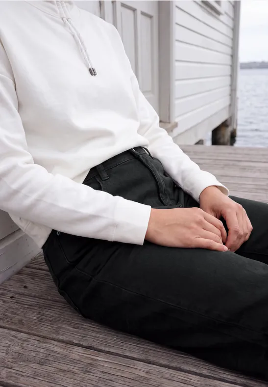 Person sitzt auf Holzsteg am Wasser, trägt weißen Pullover und schwarze Hose.