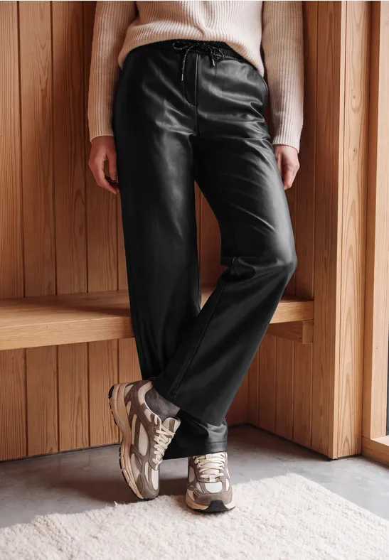 Person trägt schwarze Lederhose, hellen Pullover und Sneaker, steht vor hölzerner Wand.