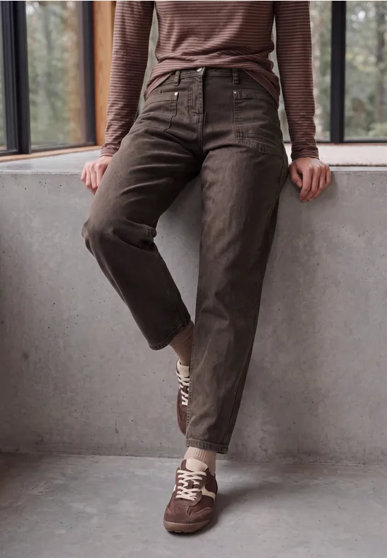Dunkelbraune, hoch taillierte Cordhose und passende Sneaker, Person lehnt an einer Betonbrüstung.