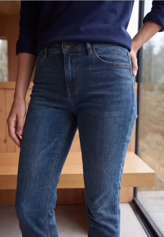 Person trägt hoch geschnittene dunkelblaue Jeans, steht neben einem Fenster vor Holzwand.
