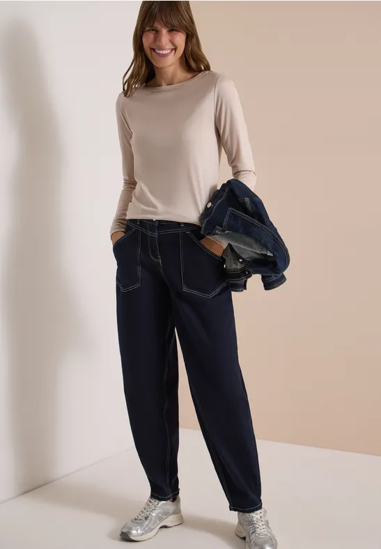 Frau in beige Langarmshirt, weiten Jeanshosen und Sneakers hält eine Jeansjacke über der Schulter.