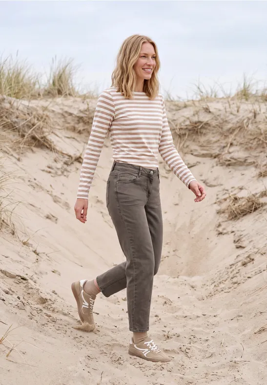 Frau in gestreiftem Pullover und dunkler Hose spaziert lächelnd über Sanddünen am Strand.