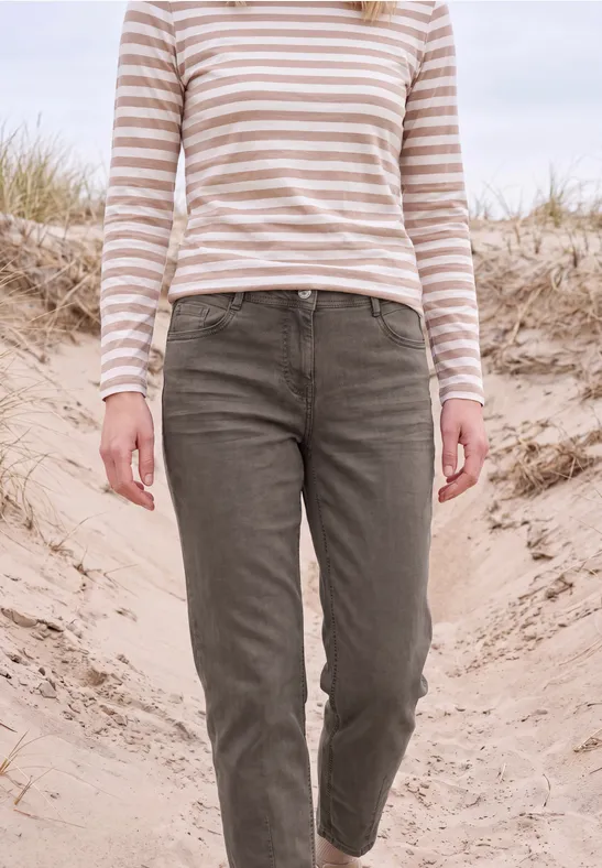 Person in beigeweiß gestreiftem Shirt und olivgrünen Hosen läuft über Sanddünen, Hintergrund mit Strandgras.