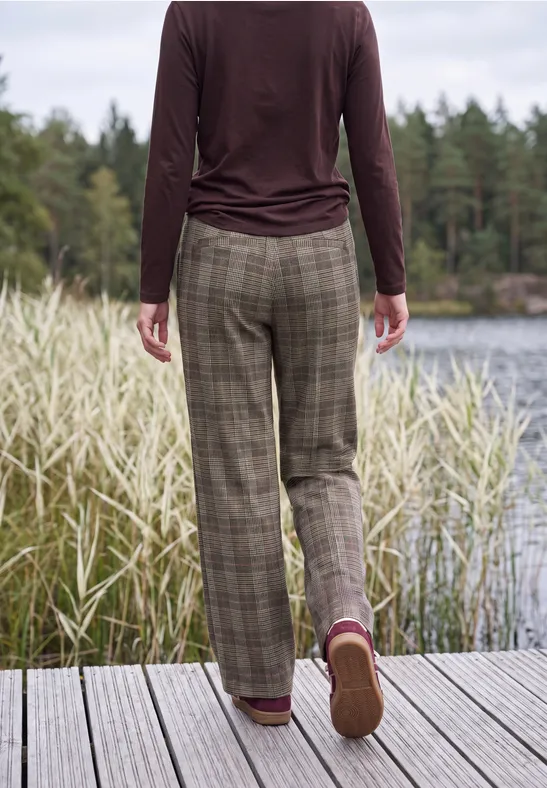 Person in karierter Hose und Langarmshirt steht auf einem Steg am See, umgeben von Schilf und Wald.