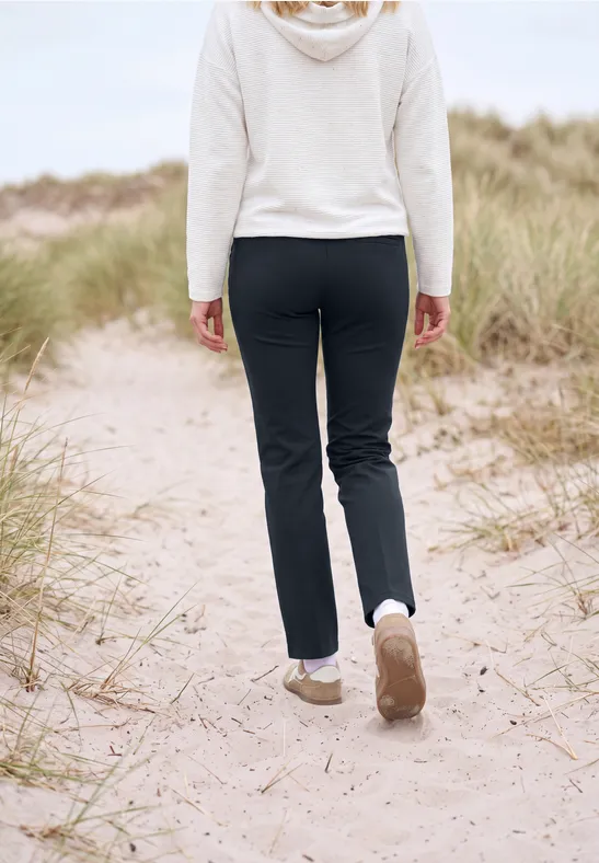 Une personne en pantalon noir et pull blanc marche sur un sentier de dunes sableuses avec herbes.