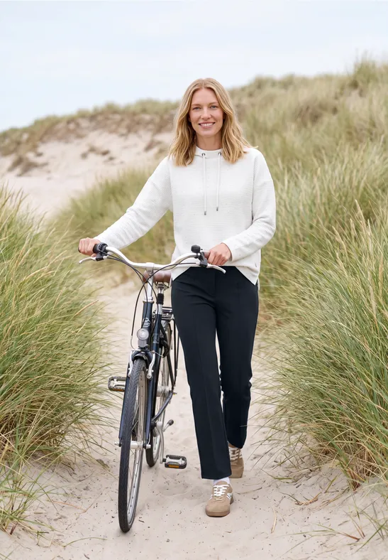 Femme souriante pousse son vélo dans les dunes près de la plage, en pull blanc et pantalon noir.