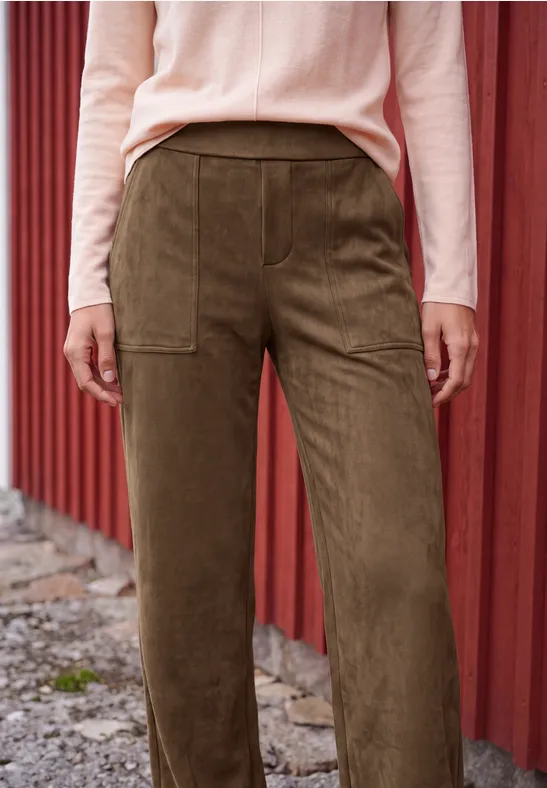 Damenhose aus grünem Cord mit großen Taschen, kombiniert mit rosafarbenem Langarmpullover vor roter Holzwand.