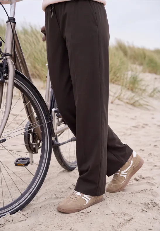 Osoba w czarnych luźnych spodniach i beżowych trampkach na piaszczystej plaży obok roweru.