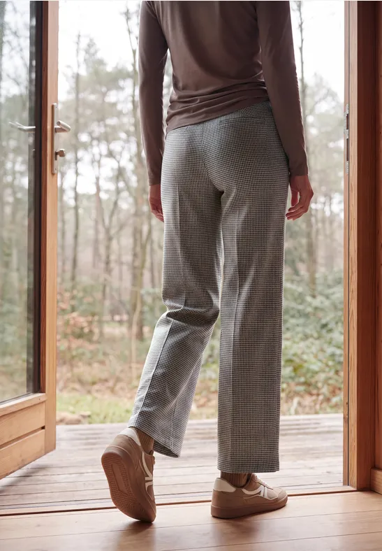 Person steht auf einer Holzterrasse vor einer Glastür, trägt graue Hose und beigen Sneaker, im Hintergrund Bäume.