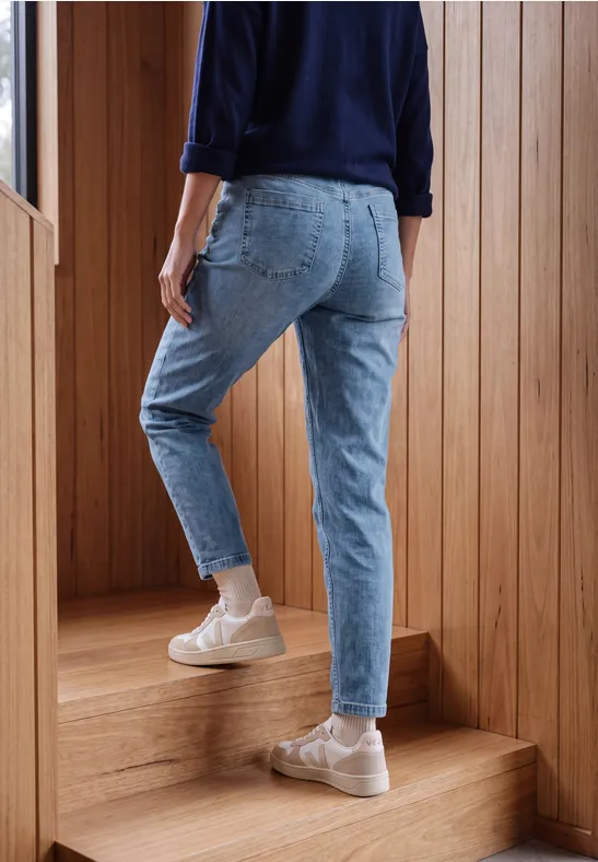 Een persoon in blauwe spijkerbroek en lichte sneakers loopt een houten trap op.