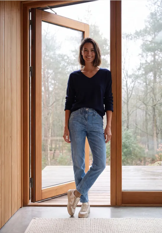 Vrouw staat glimlachend in de open deur van een houten huis, met bos en daglicht op de achtergrond.