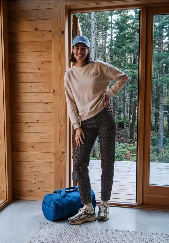 Person in Jogginghose und Cap steht in Holzhütte vor großen Fenstertüren mit Blick auf Wald, blaue Reisetasche daneben.