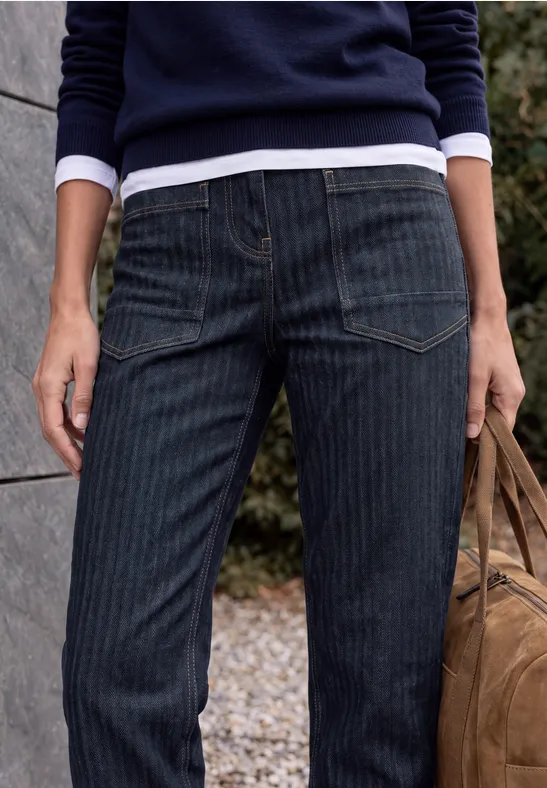 Figur in dunkler Nadelstreifenjeans, Marinepullover und Tasche, steht vor einer Mauer im Freien.