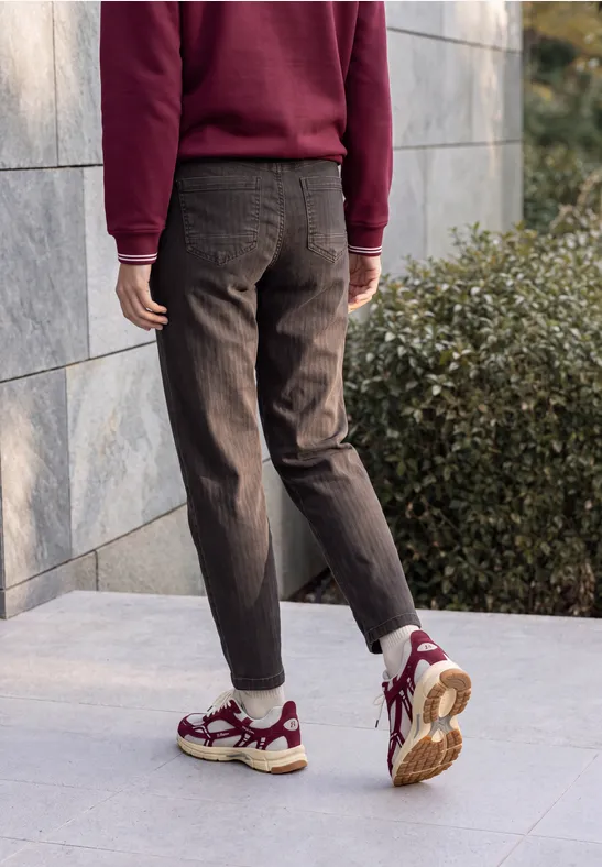 Persoon in bordeauxrood trui en donkere broek, witte sneakers met rode details, loopt langs een stenen muur.