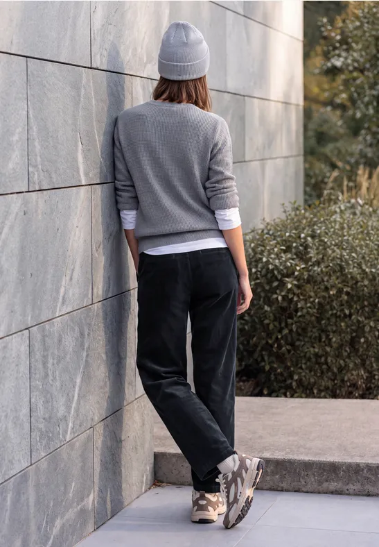 Person mit grauer Mütze und Sweatshirt lehnt an Steinmauer, trägt schwarze Hose und Sneaker.