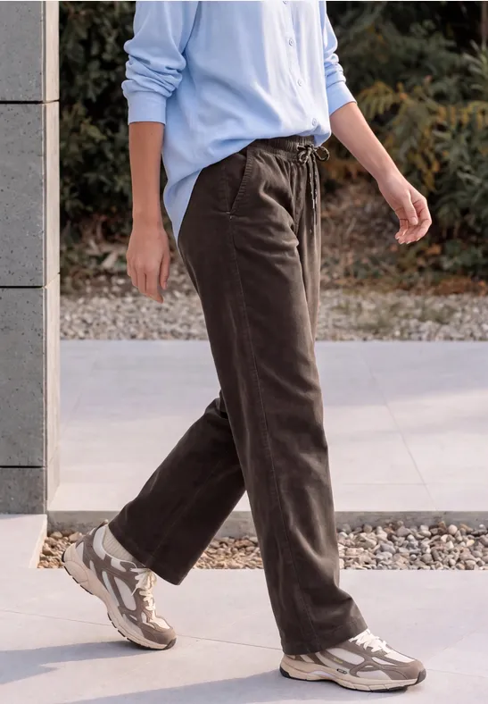 Personne en pantalon en velours côtelé marron, chemise bleu clair et baskets beige sur un trottoir.