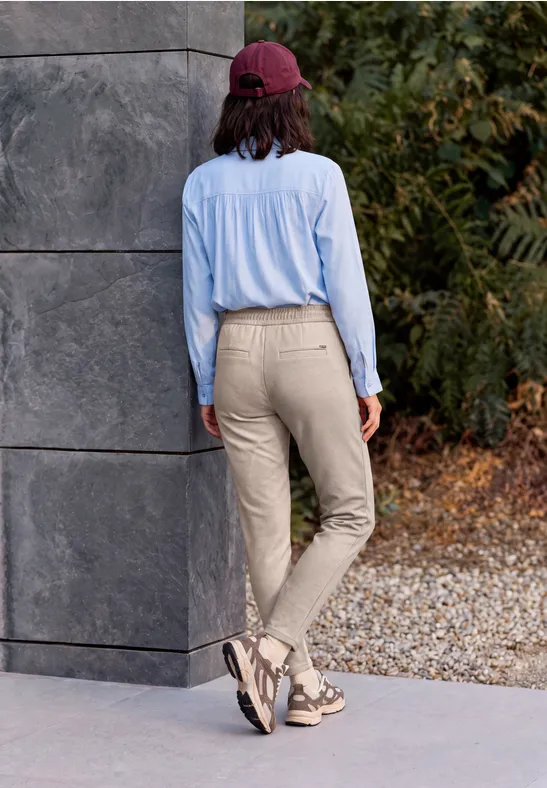 Person mit roter Kappe, hellblauer Bluse und beigen Hosen läuft an Mauer und herbstlichem Laub vorbei.