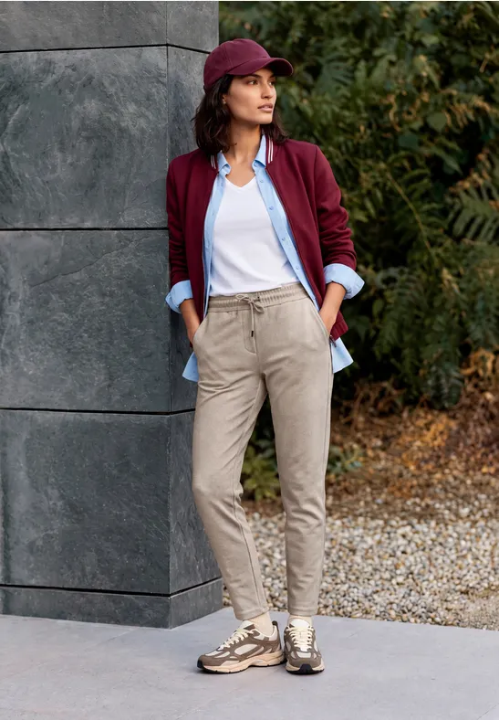 Frau lehnt an Hauswand im Freien, trägt burgunderrote Jacke, hellblaue Bluse, weißes Top, beige Hose und Sneaker.
