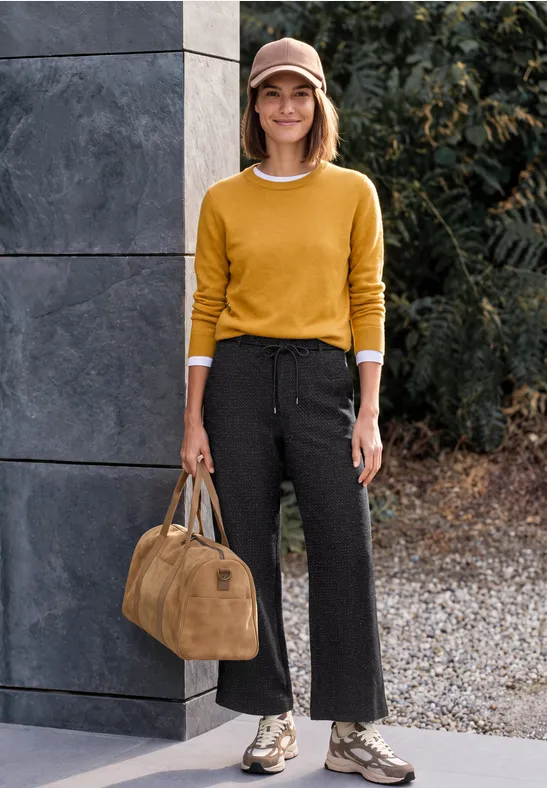 Frau mit gelbem Pullover, schwarzer weiter Hose, Schlangenprint-Stiefeln und Tasche steht vor moderner Fassade im Freien.