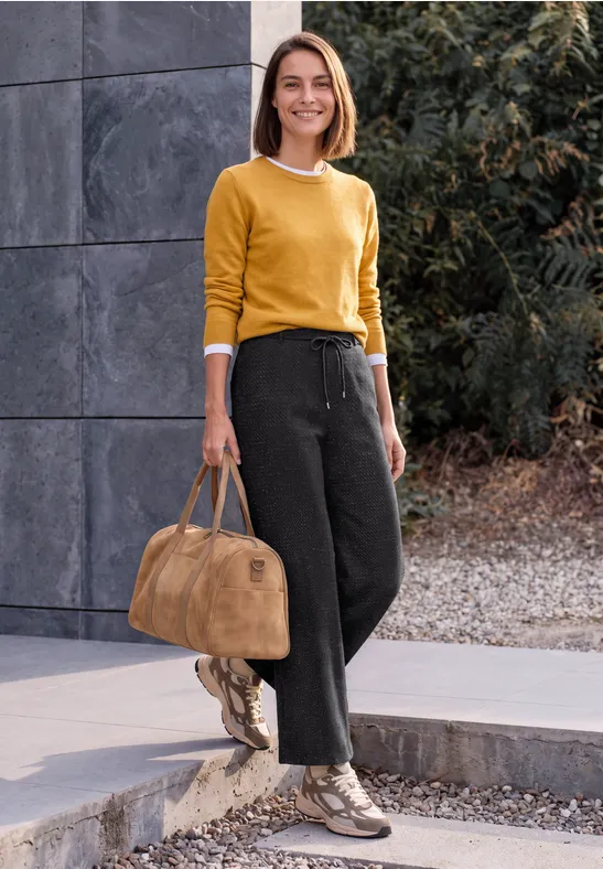Frau in gelbem Pullover und schwarzer Hose mit beiger Tasche, draußen stehend vor einer grauen Betonwand und Grün.