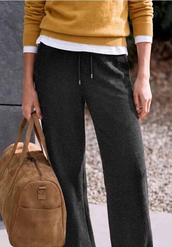 Eine Person trägt schwarze Hose und gelben Pullover und hält eine braune Wildledertasche.