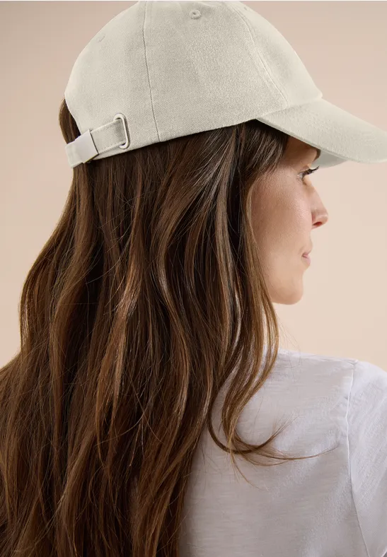 Frau von hinten mit langen braunen Haaren, weißem Shirt und beiger Baseballkappe vor beigem Hintergrund.