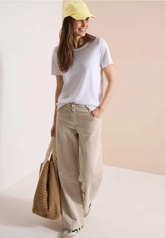 Frau in weißem T-Shirt, weitem beigen Stoffhosen und gelber Mütze mit Strohtasche.