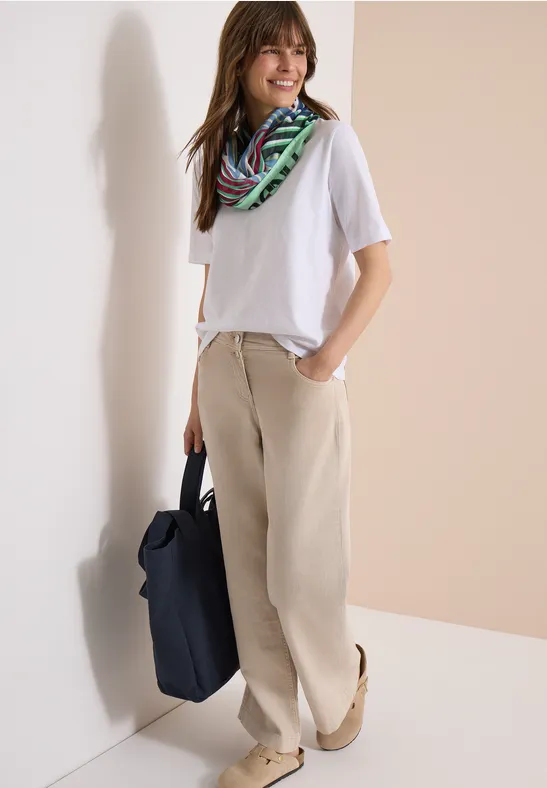 Frau lehnt an Wand, trägt weißes T-Shirt, bunten Schal, beige Stoffhose und hält eine schwarze Tasche.