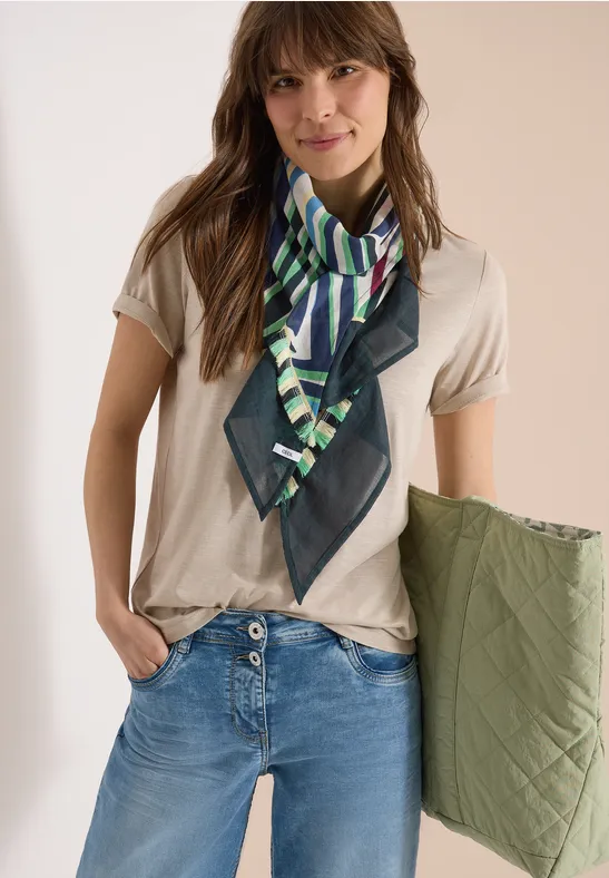 Frau in beigem T-Shirt, blauer Jeans, grün gemustertem Seidenschal, hält olivgrüne gesteppte Jacke
