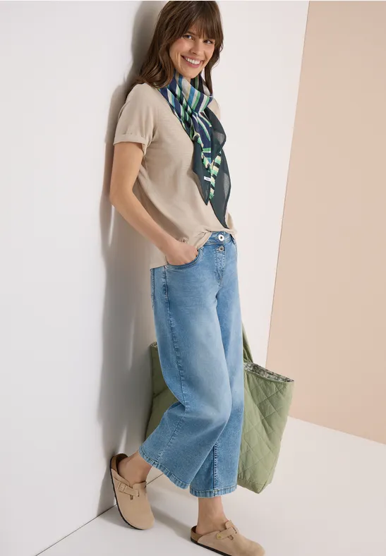 Eine Frau lehnt an einer Wand, trägt weite Jeans, ein beiges Top, grünen Schal und grüne Tasche.