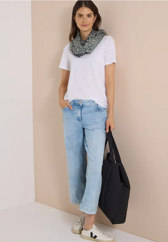 Frau in weißem T-Shirt, hellem Jeans, gemustertem Loop-Schal, hält große schwarze Tasche vor beigem Hintergrund.