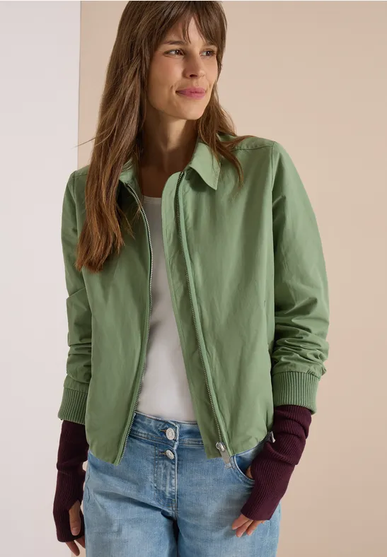 Frau trägt hellgrüne Jacke, weißes Shirt, blaue Jeans und lila Handstulpen.