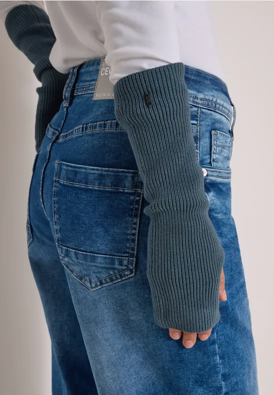 Eine Person trägt graue Strick-Handstulpen und Jeans, dazu ein weißer Pullover.
