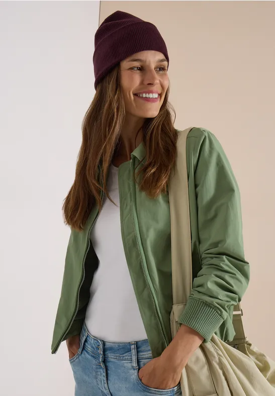 Femme souriante en veste verte, débardeur blanc, jean, bonnet violet et sac à bandoulière sur fond clair.
